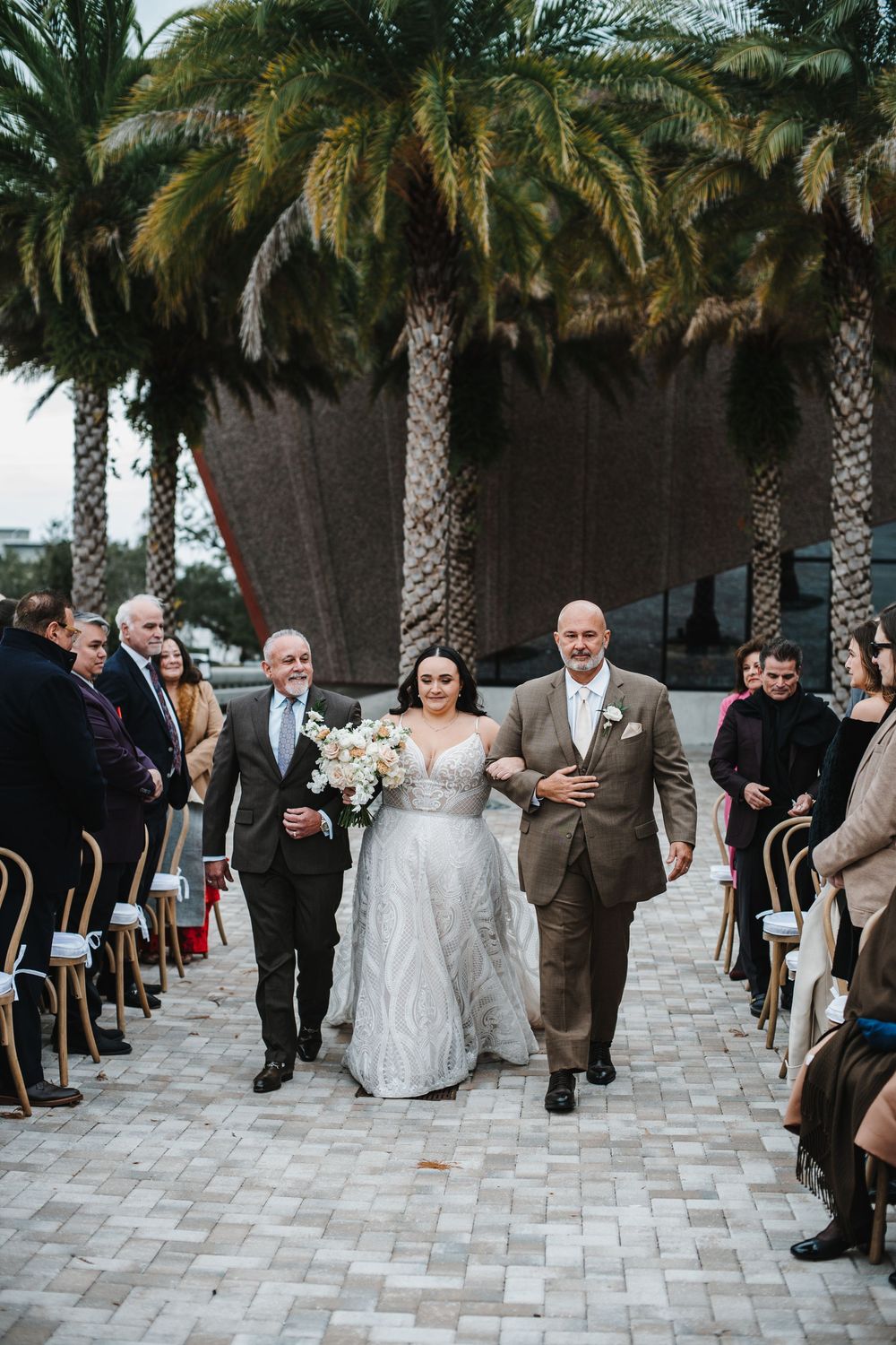 A wedding ceremony takes place outdoors under palm trees with guests seated along a stone paved aisle.