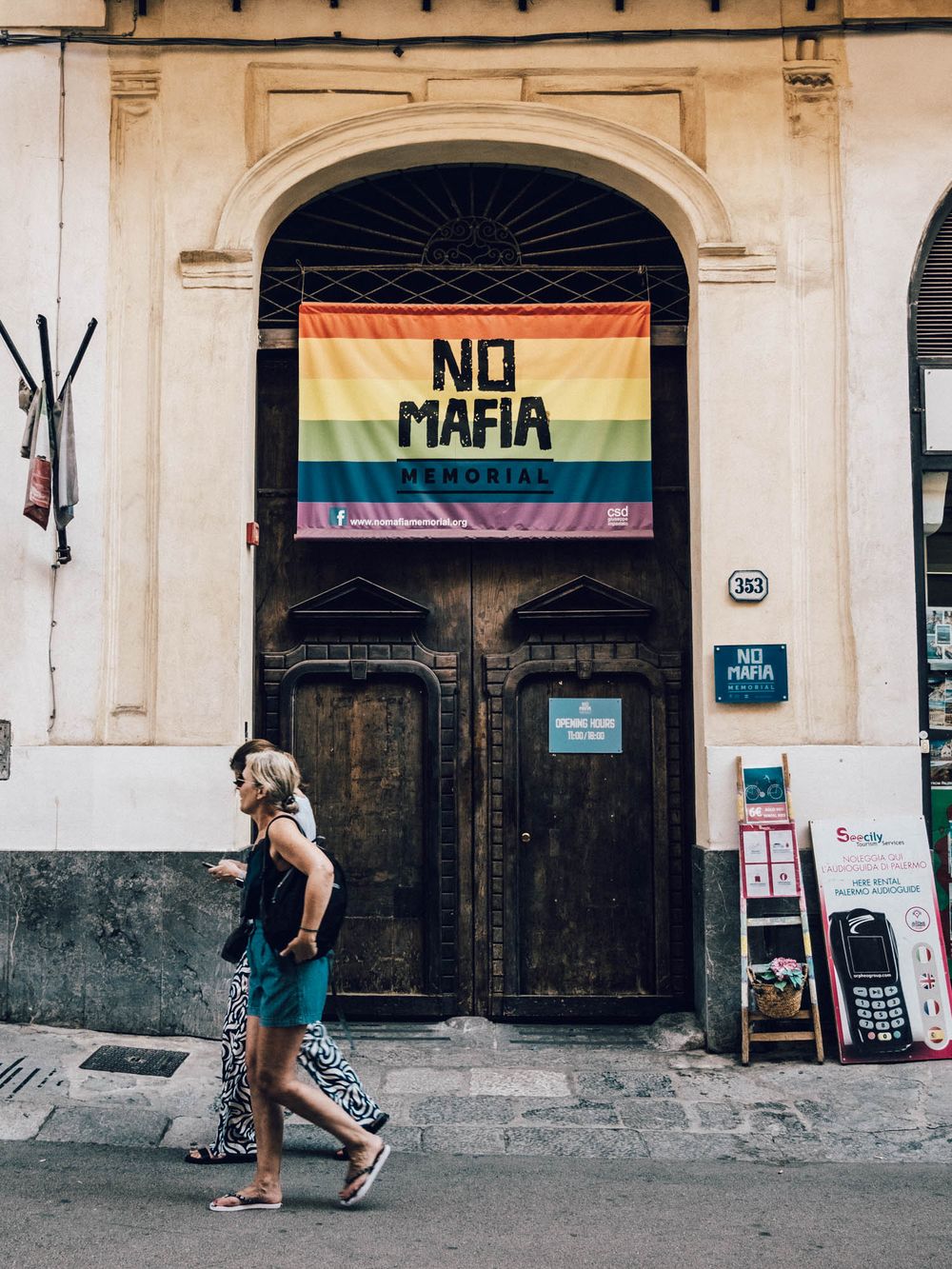 Pride flags and protest signs adorn the exterior of a historic stone building with arched doorways in an urban setting.