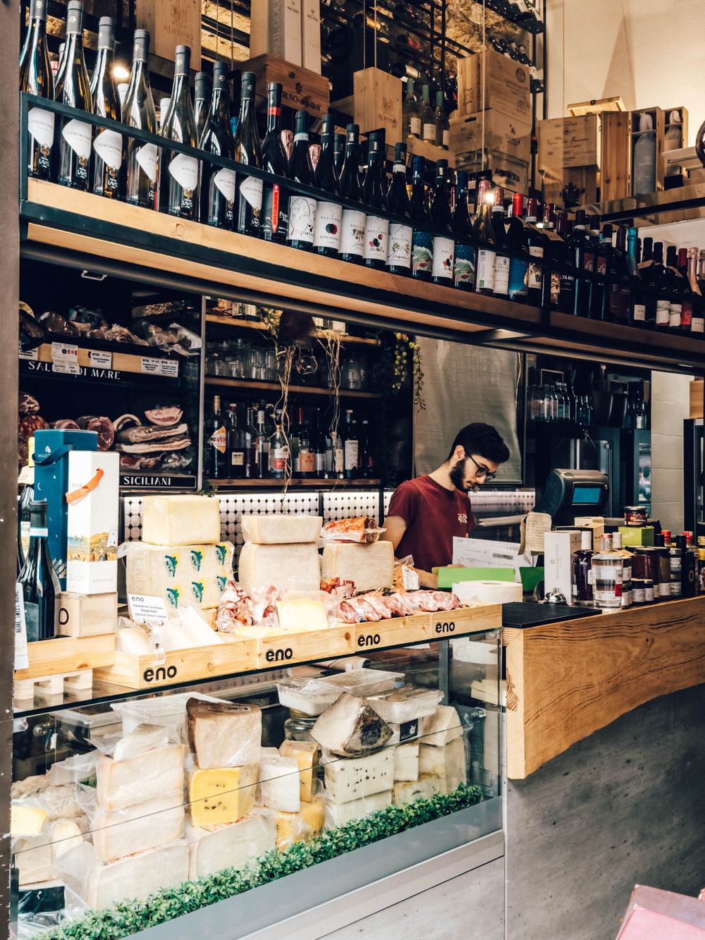A panoramic view of a gourmet market counter displaying wine bottles and artisanal cheeses on wooden shelves.