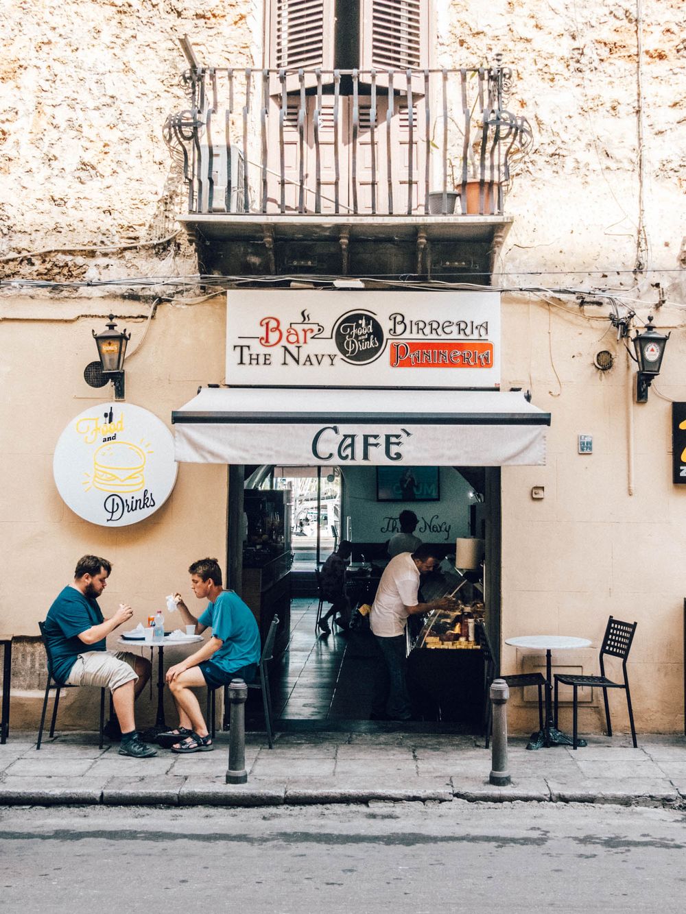 Quaint cafe storefront in a rustic building with outdoor seating and people dining at small tables on the sidewalk.