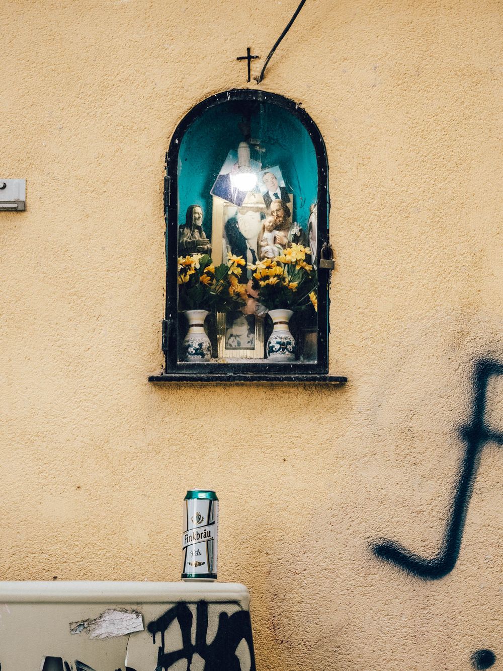 Small religious shrine with turquoise alcoves mounted on a beige stucco wall displays decorative crosses and figurines.
