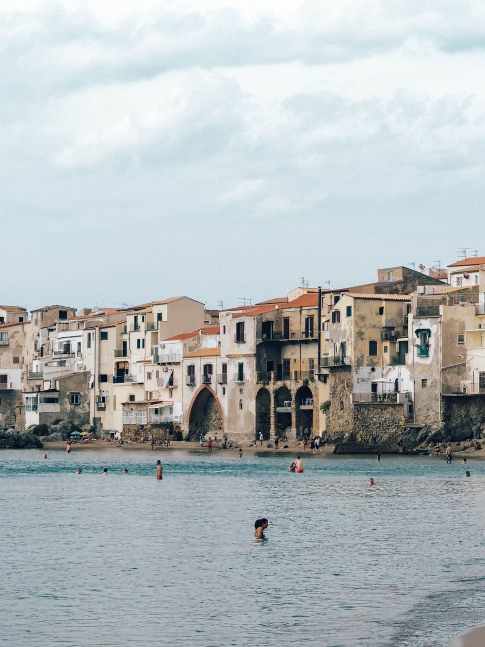 Historic waterfront houses line the Mediterranean coast of Cefalu, Sicily with boats floating in the turquoise waters.