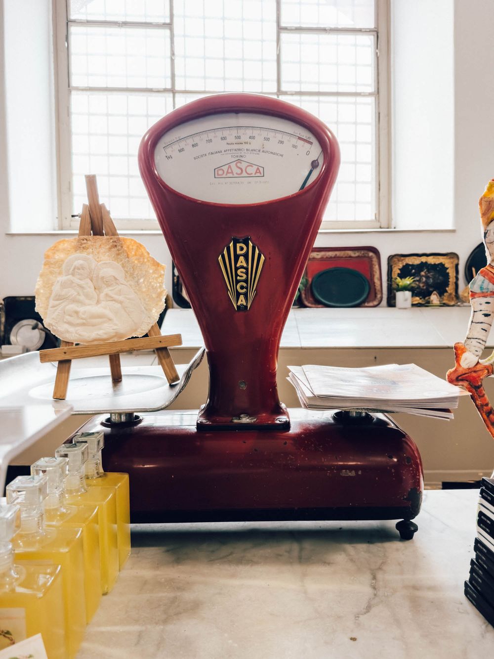 Vintage burgundy weighing scale with glass display window sits on counter next to yellow bottles in bright interior.