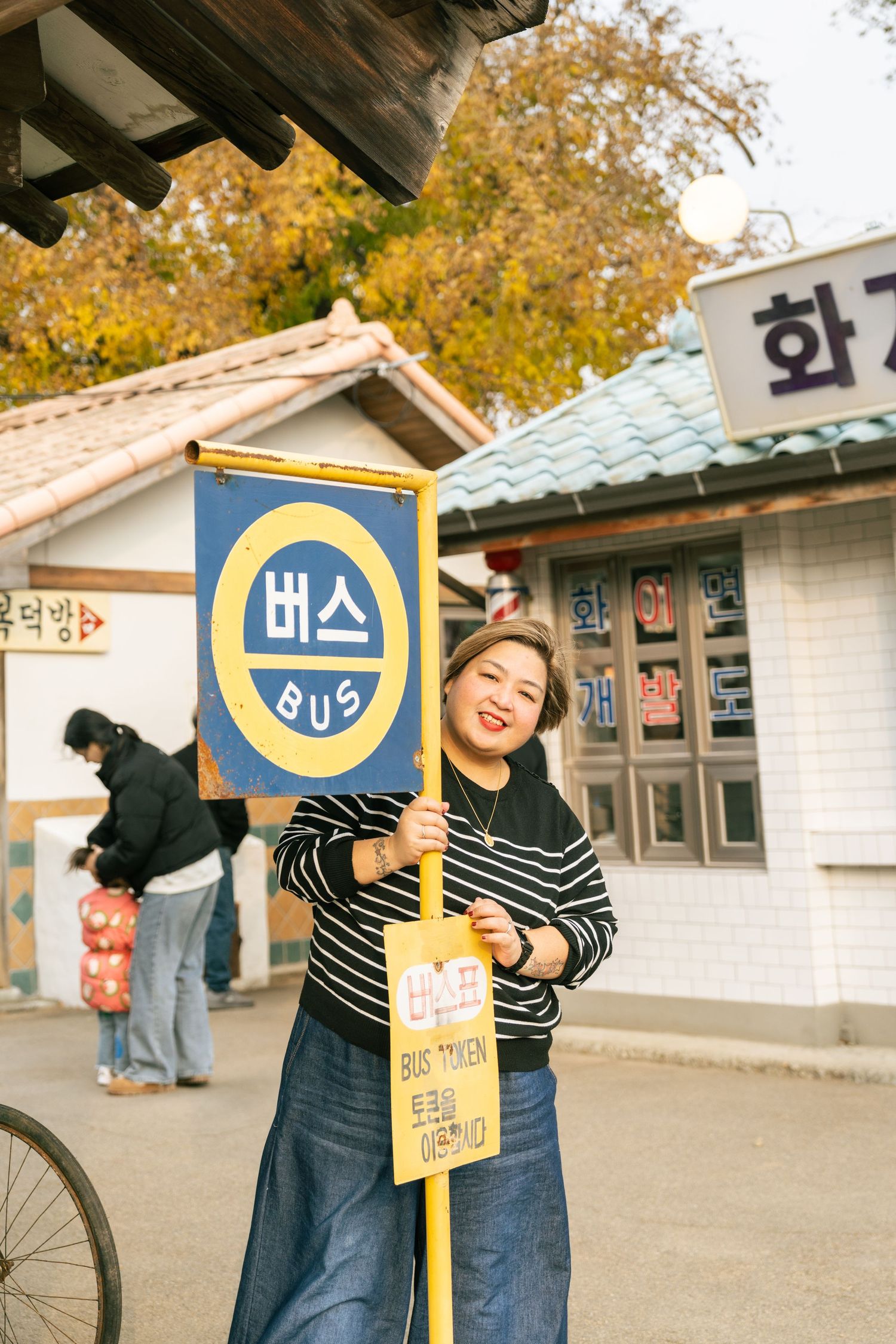 A person in a striped shirt poses playfully with a yellow and blue bus stop sign at a traditional Korean village.