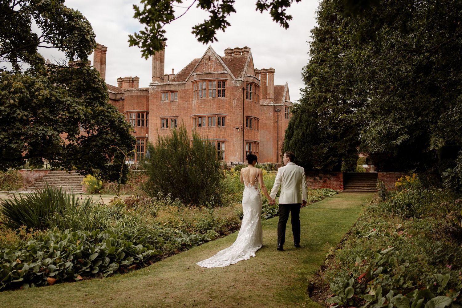 A bride and groom walk together through a lush garden path at an elegant historic brick estate.