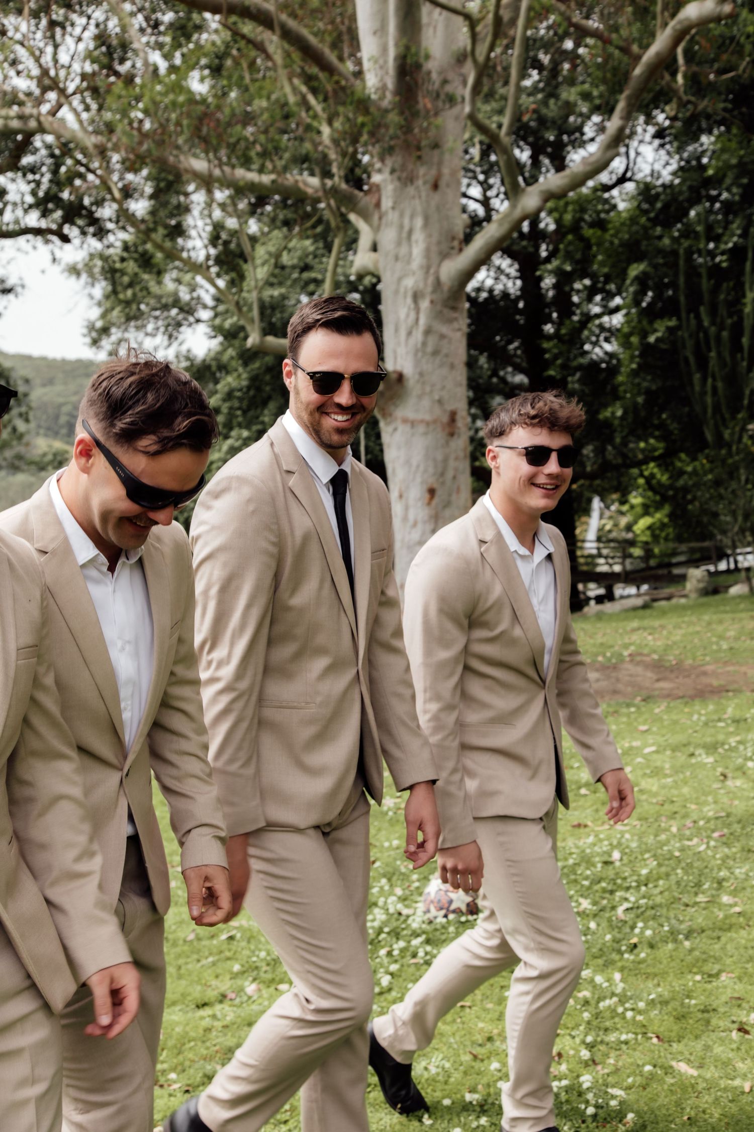 Groomsmen in beige suits and sunglasses walking in line on grass with white flowers in a playful manner.
