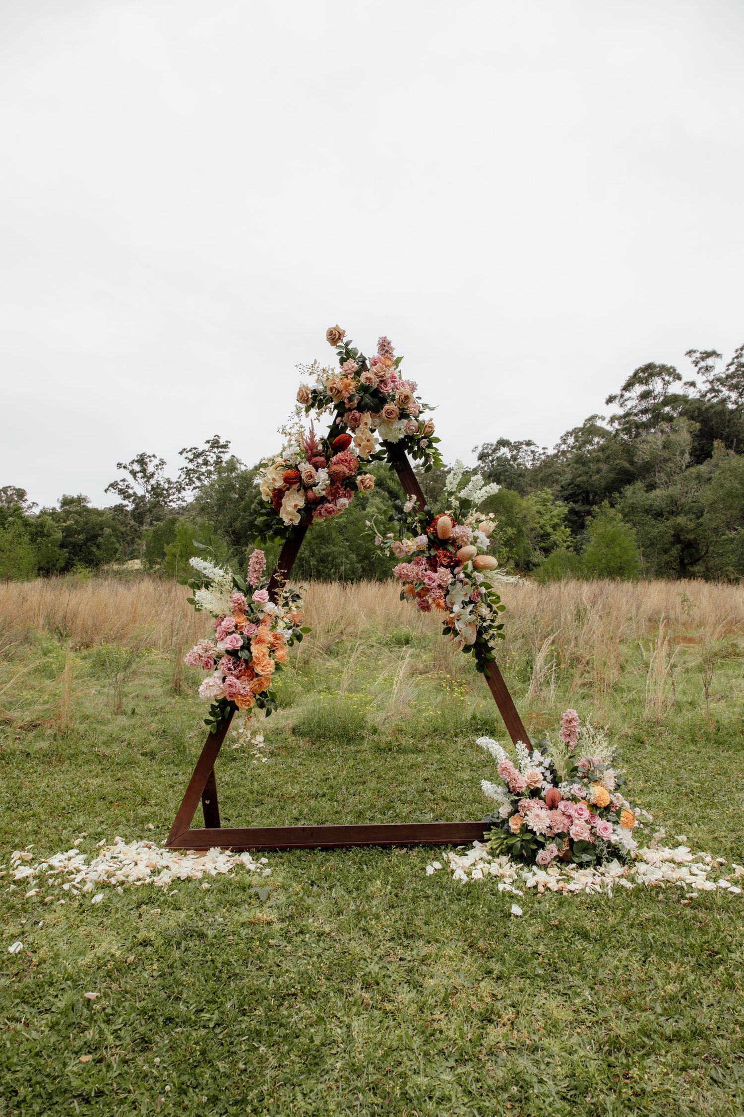 Rustic wooden arch decorated with pink and peach flowers stands in a grassy meadow with scattered flower petals below.