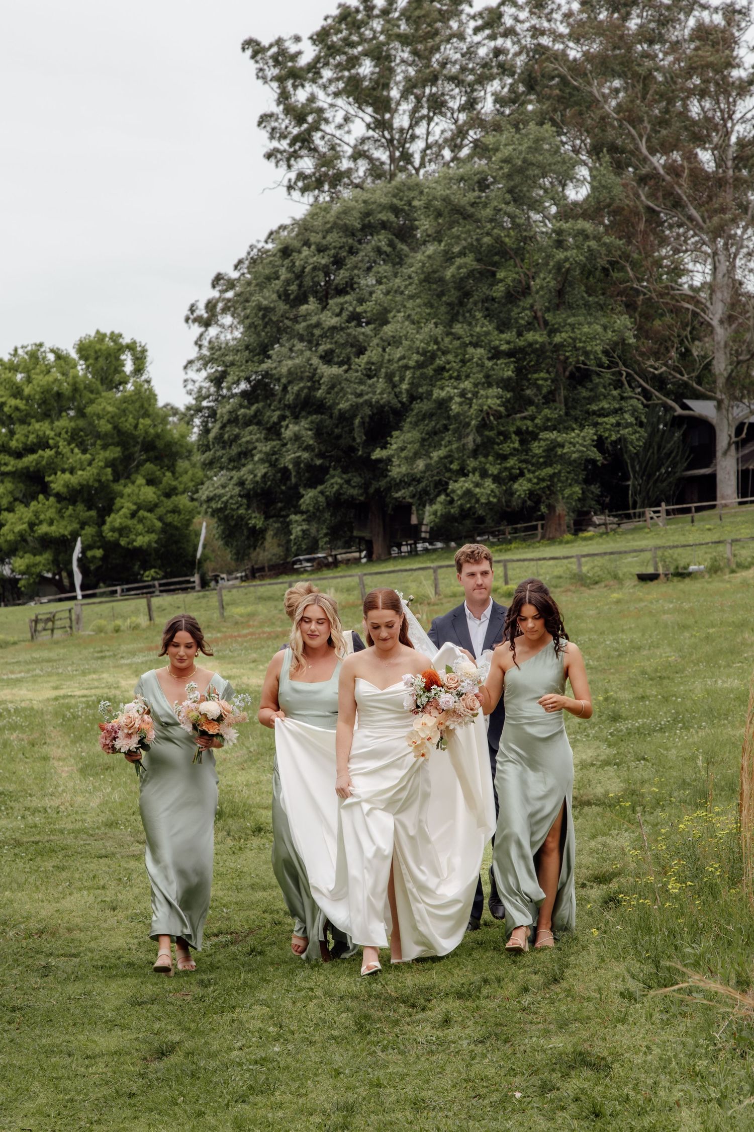Bridesmaids in sage green dresses walk together across a grassy hill at an outdoor wedding venue.