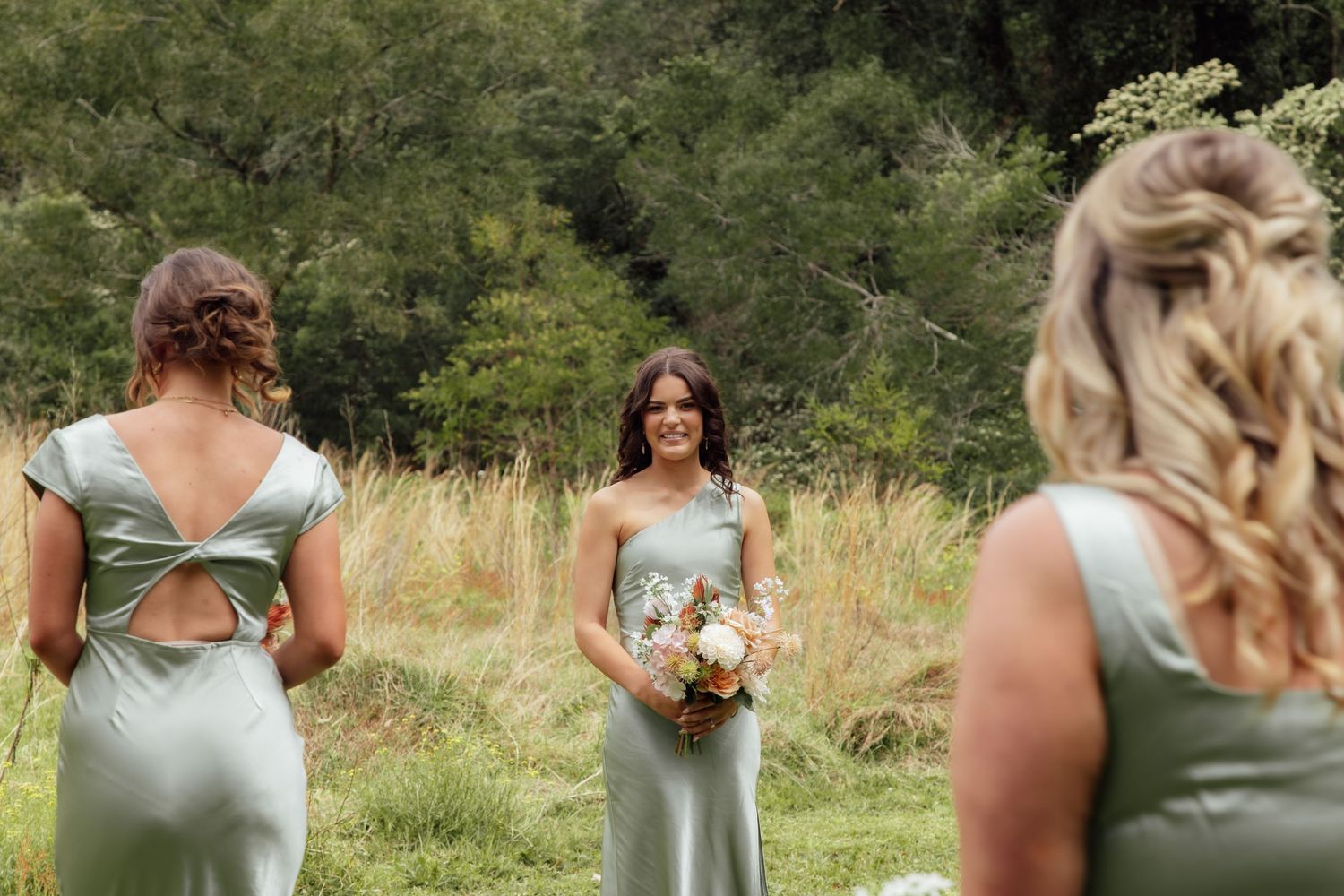 A row of bridesmaids in sage green dresses stand together holding bouquets in an outdoor garden setting.
