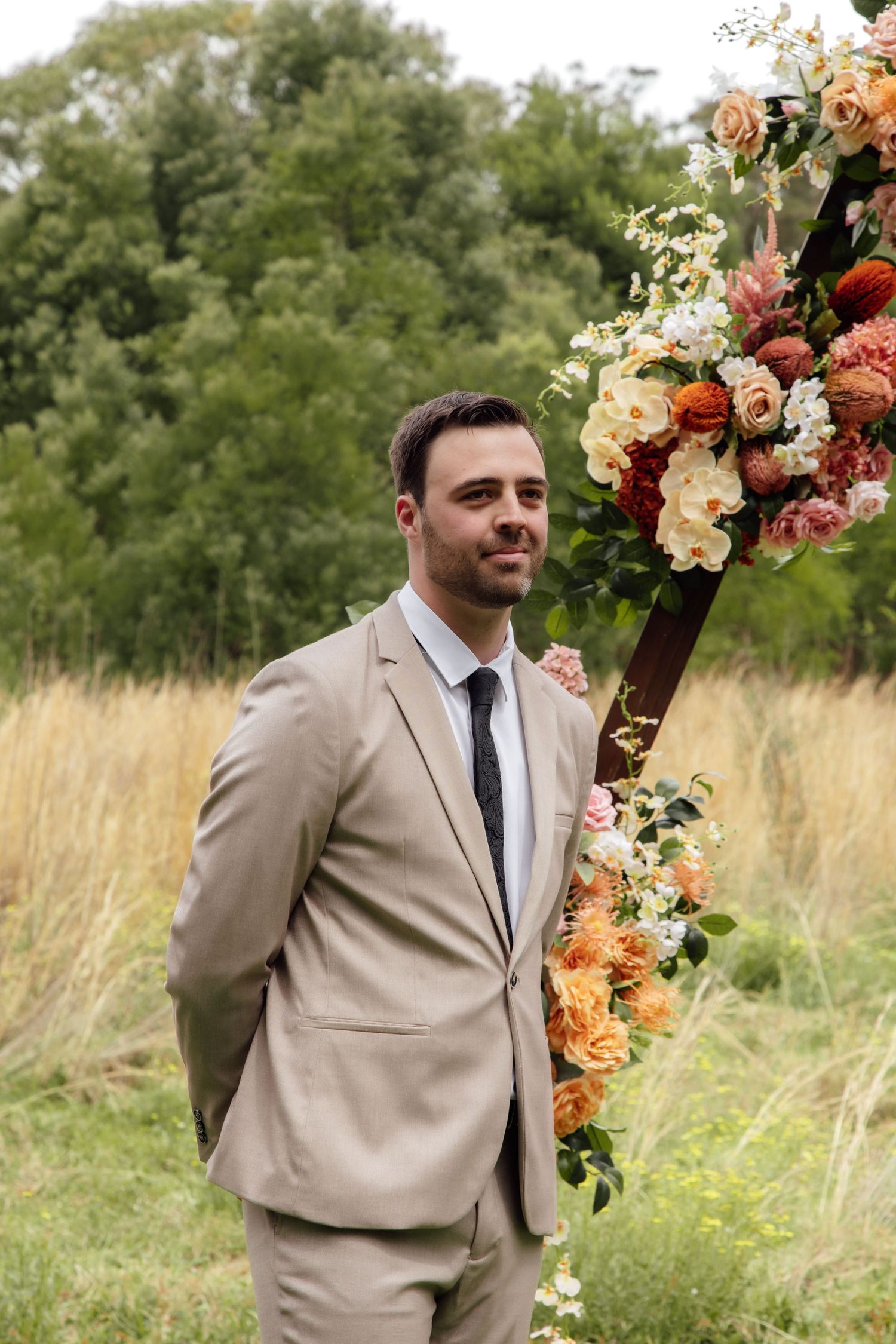 A floral arch with peach and coral blooms frames a scenic outdoor setting with tall grass and trees in the background.
