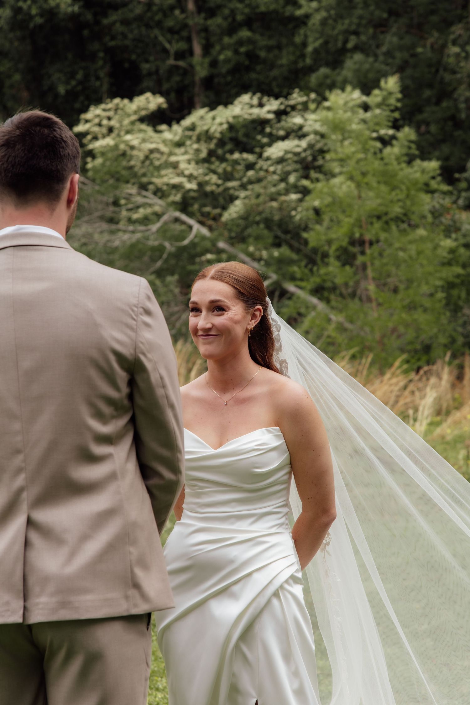 A bride in a white strapless gown smiles joyfully while walking outdoors with her partner in a tan suit.