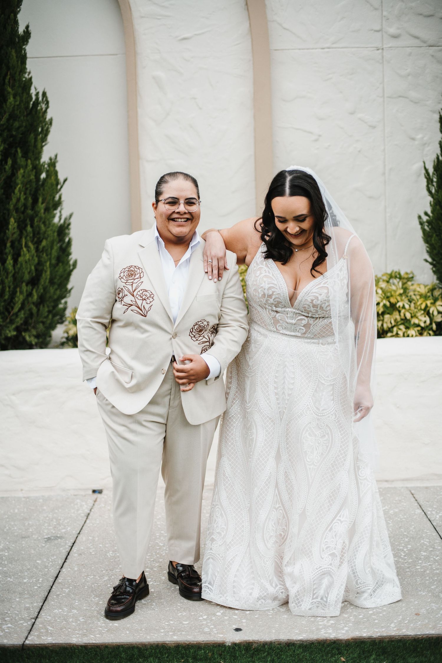A couple in coordinated cream-colored wedding attire stand together smiling outdoors against a white wall.
