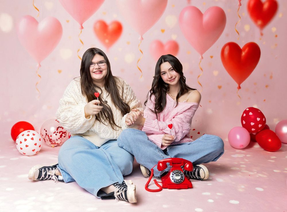 Two teen best friends sit on pink Valentine’s set holding heart props with red balloons and vintage phone in New Hampshire studio