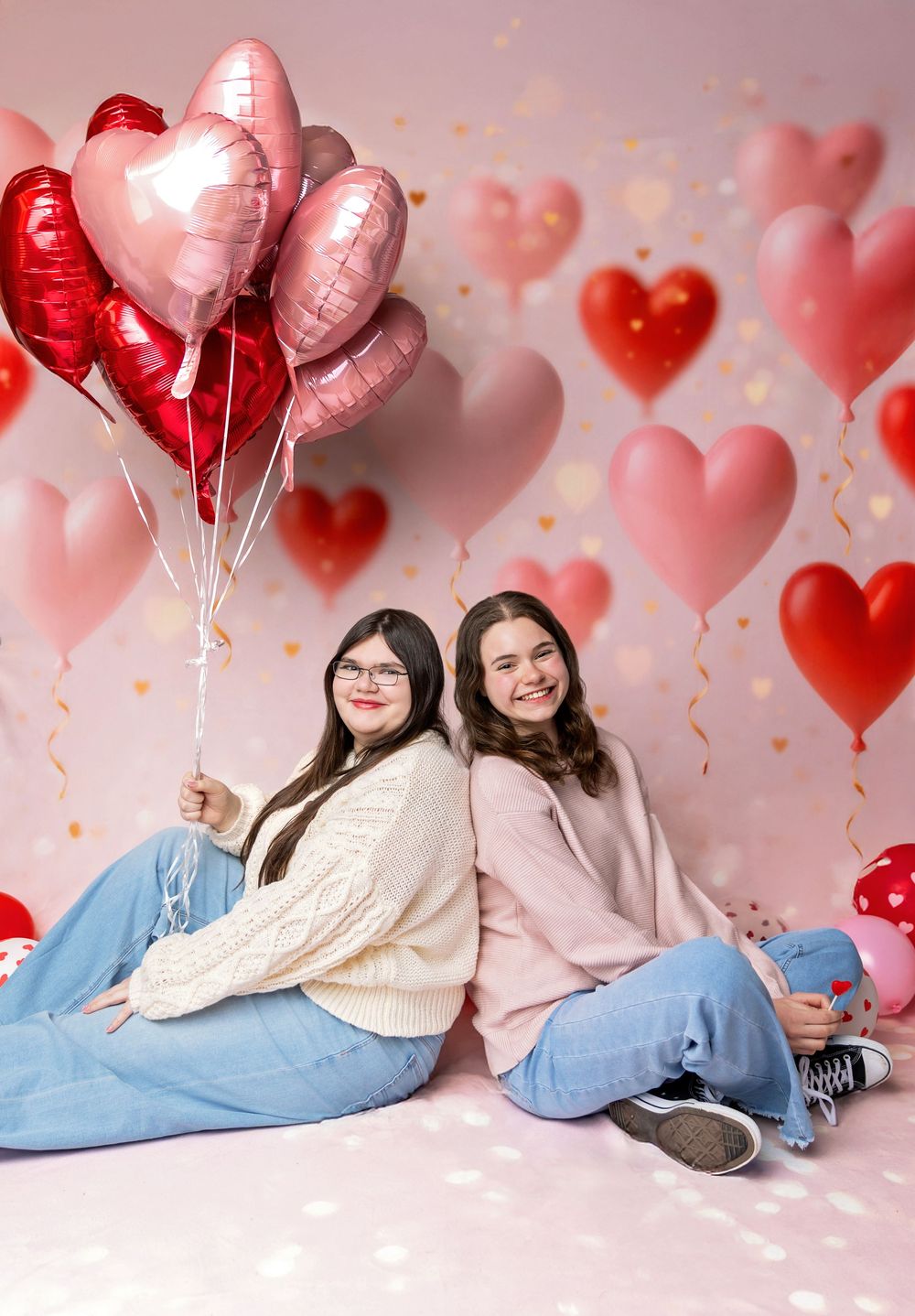 Teen best friends sit back to back holding heart balloons on pink Valentine’s backdrop in New Hampshire studio