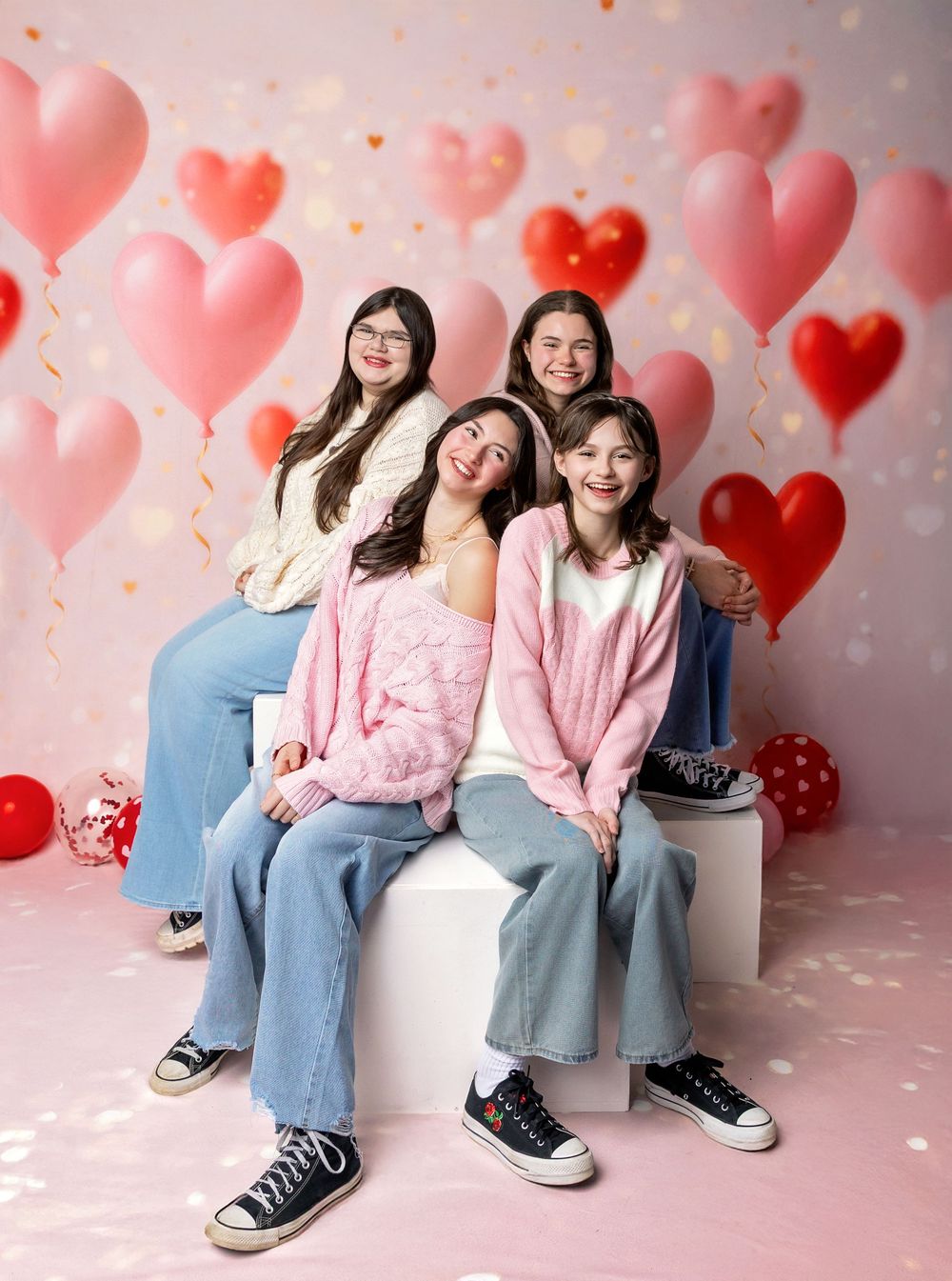 Teen girls sit with heart balloons on Valentine’s Day set with pink hearts in New Hampshire photography studio