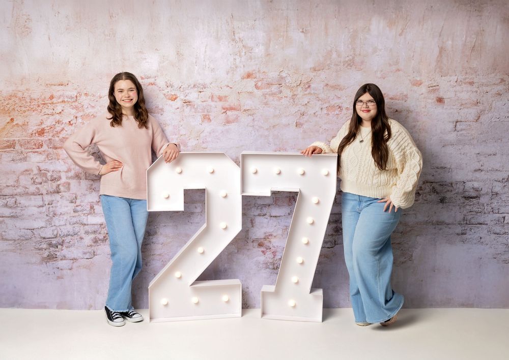 Two Class of 2027 teen girls pose with marquee 27 numbers on textured pink wall in New Hampshire studio