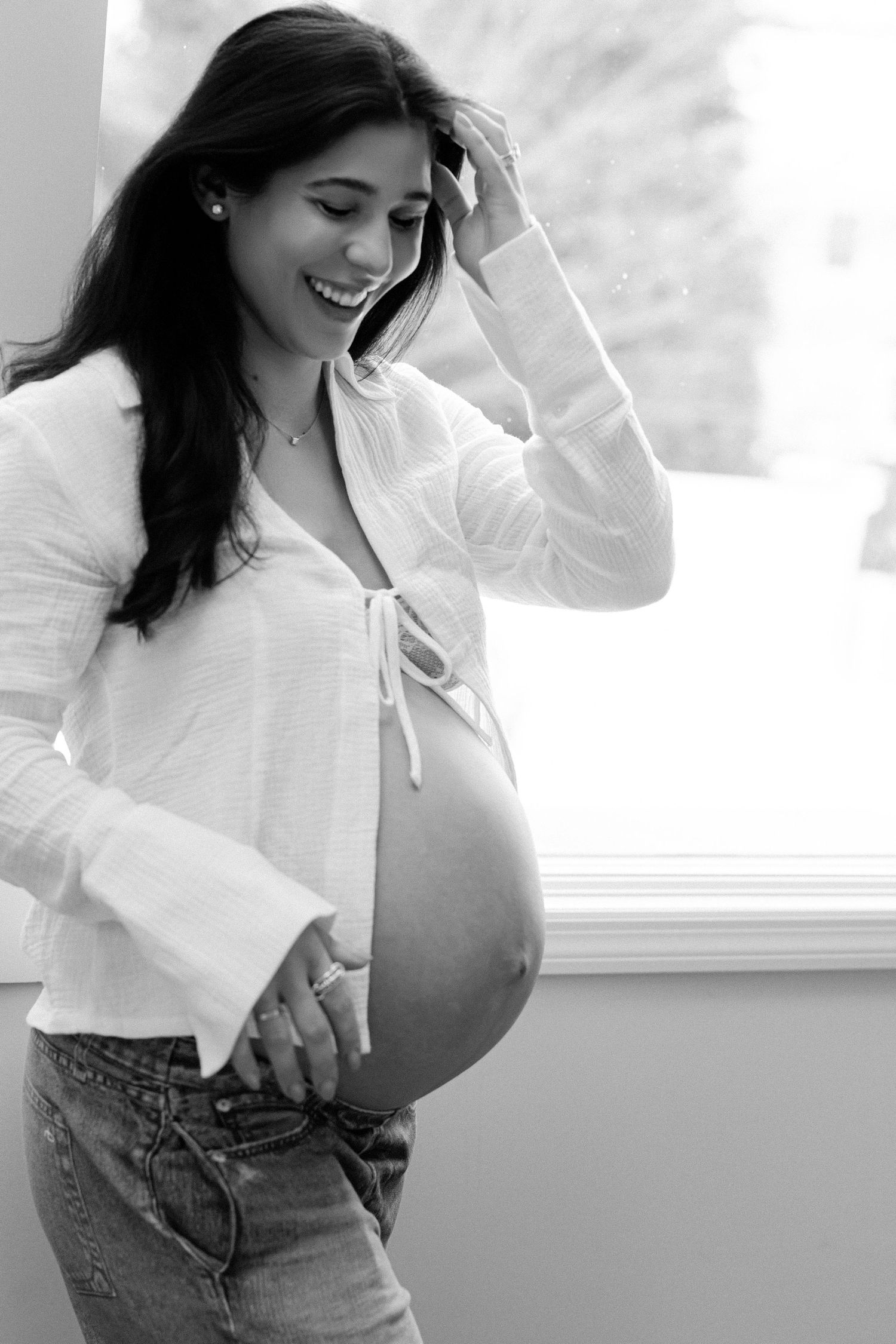 Smiling pregnant woman in white top and jeans cradling her bare baby bump near a bright window.