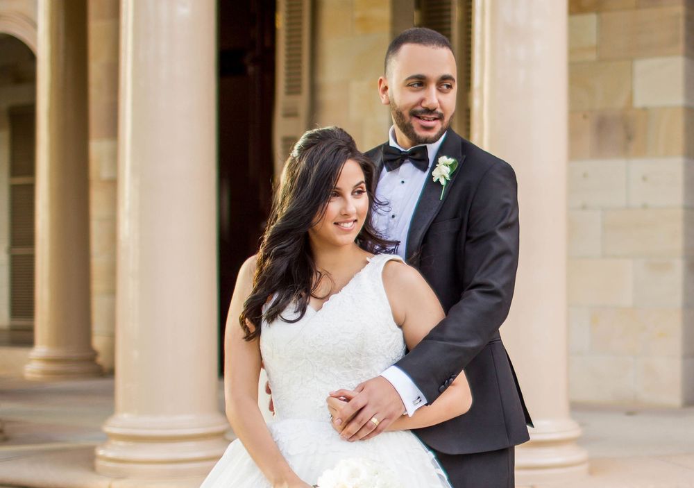 Smiling wedding couple embracing before sandstone columns, groom in dark suit, bride in white lace gown.