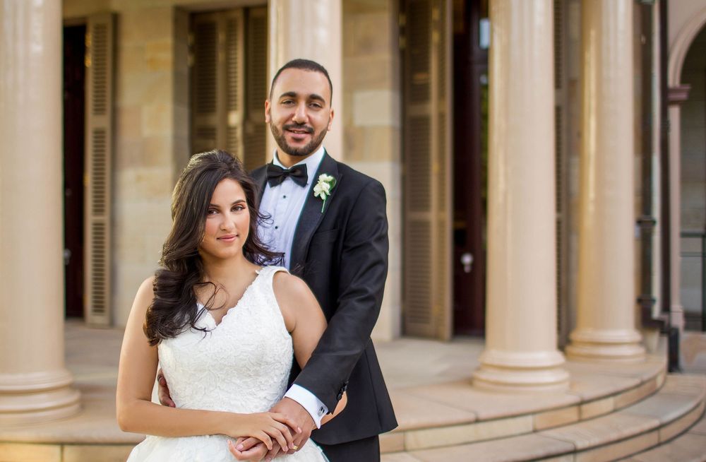 Wedding couple posing closely together before elegant sandstone columns, groom embracing bride from behind.