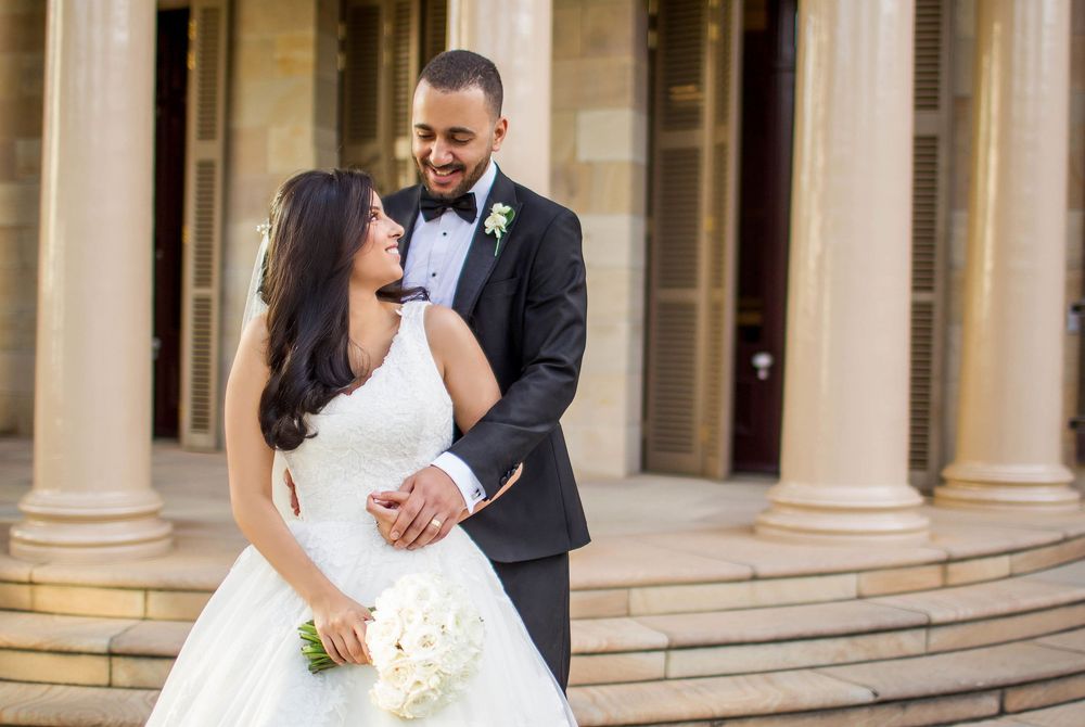 Joyful bride and groom sharing a laugh on heritage building steps, bride holding white bouquet.