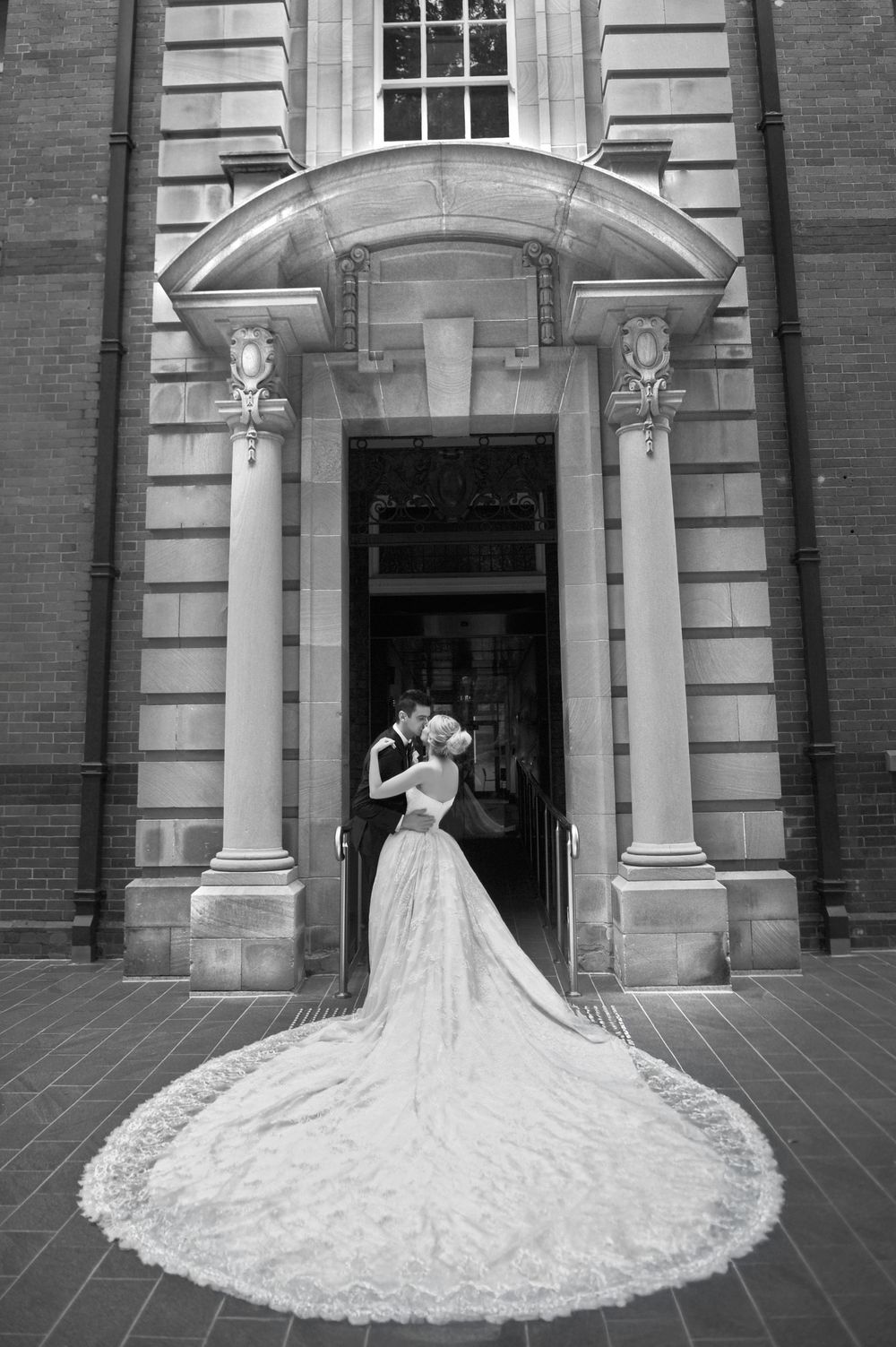 Black and white photo of bride and groom kissing in doorway of historic brick building with lace cathedral train.