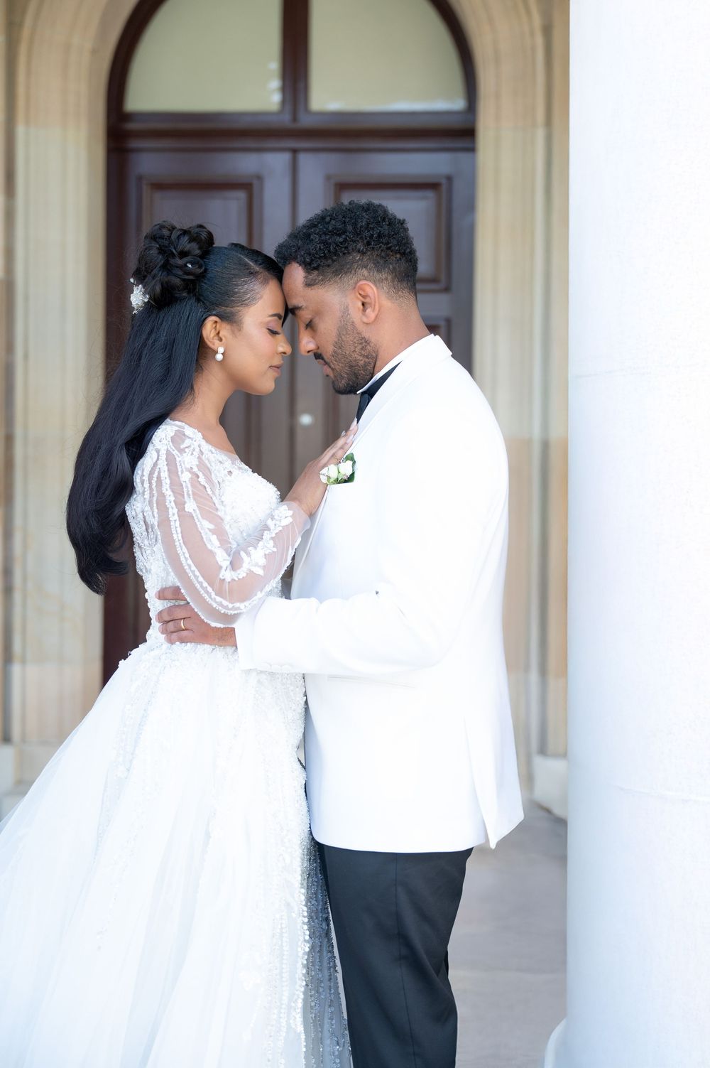 Elegant bride and groom in white wedding attire sharing intimate moment before ornate wooden doors.