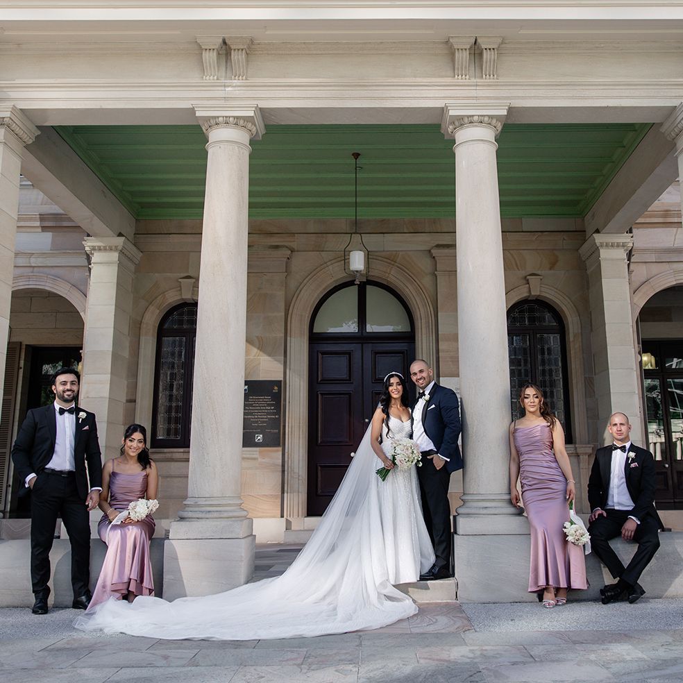 Elegant wedding party in formal attire poses on steps of a neoclassical building with white columns.