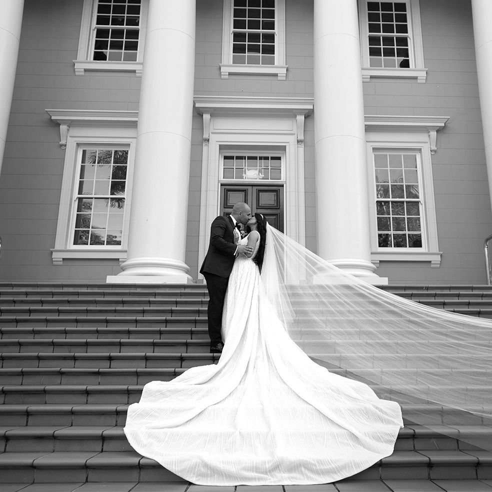 Black and white wedding portrait of couple kissing on steps of classical building with dramatic flowing veil.