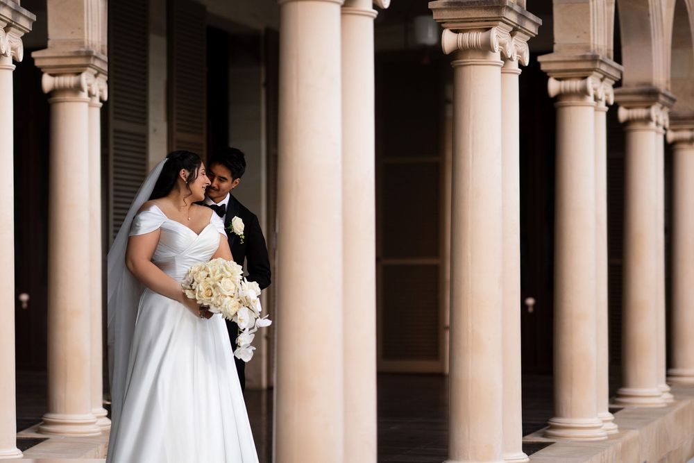 Bride holding white bouquet and groom sharing tender moment between elegant stone columns.