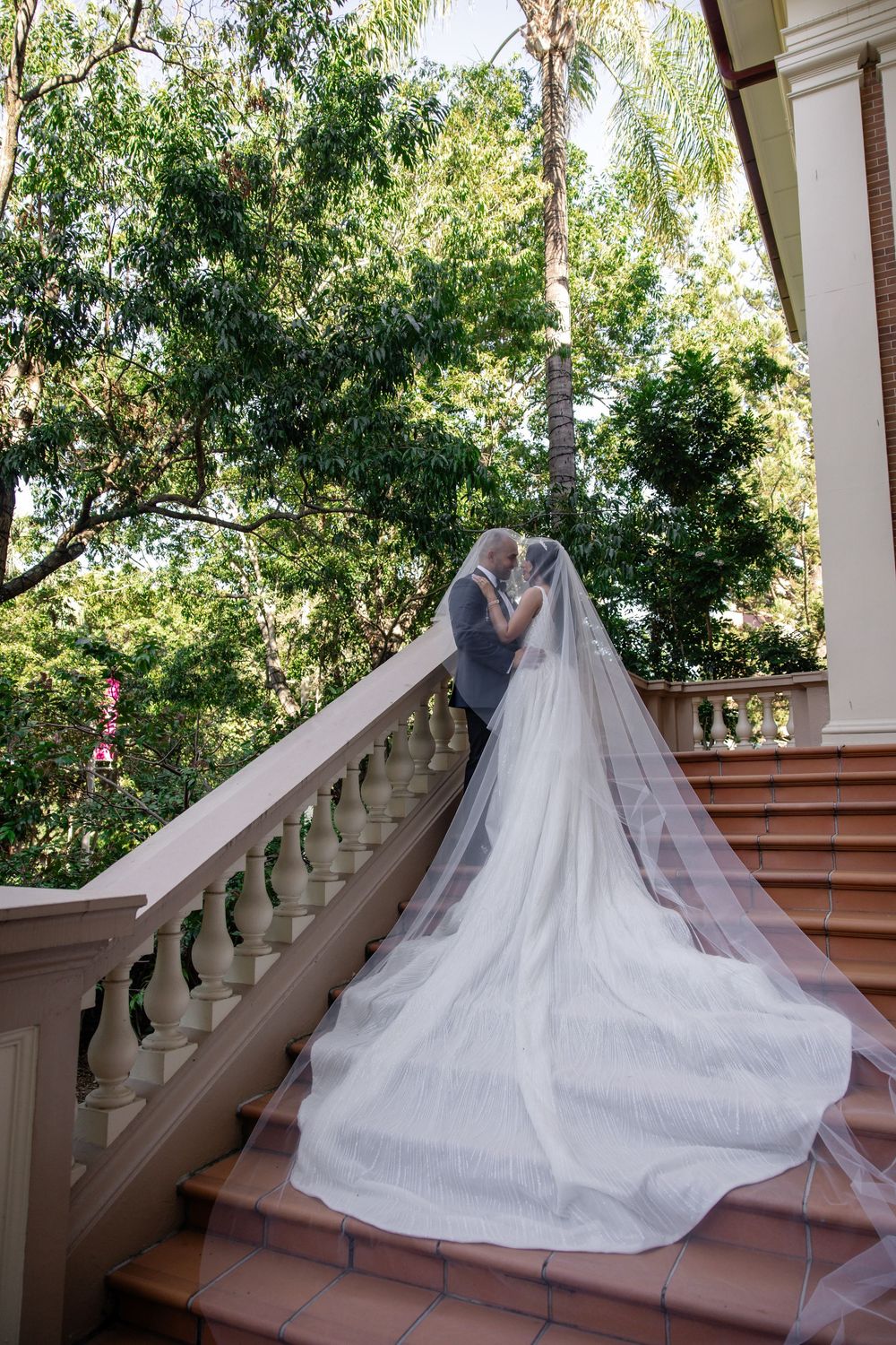 Bride and groom share a romantic kiss under a flowing veil on elegant stone stairs surrounded by lush greenery.