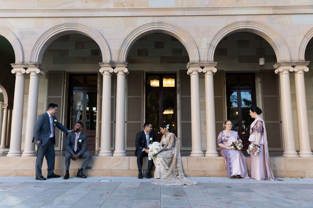 Wedding party relaxes near ornate stone arched colonnade as bride and groom share a quiet intimate moment.