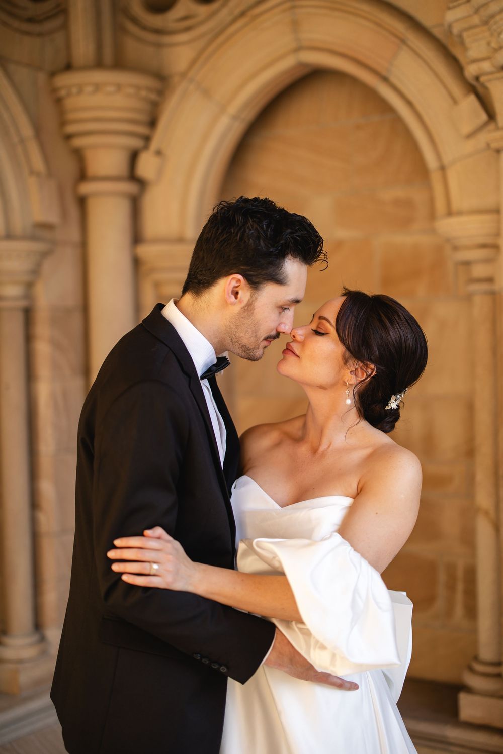 Bride and groom share a tender almost-kiss embrace framed by a stunning ornate sandstone archway backdrop.