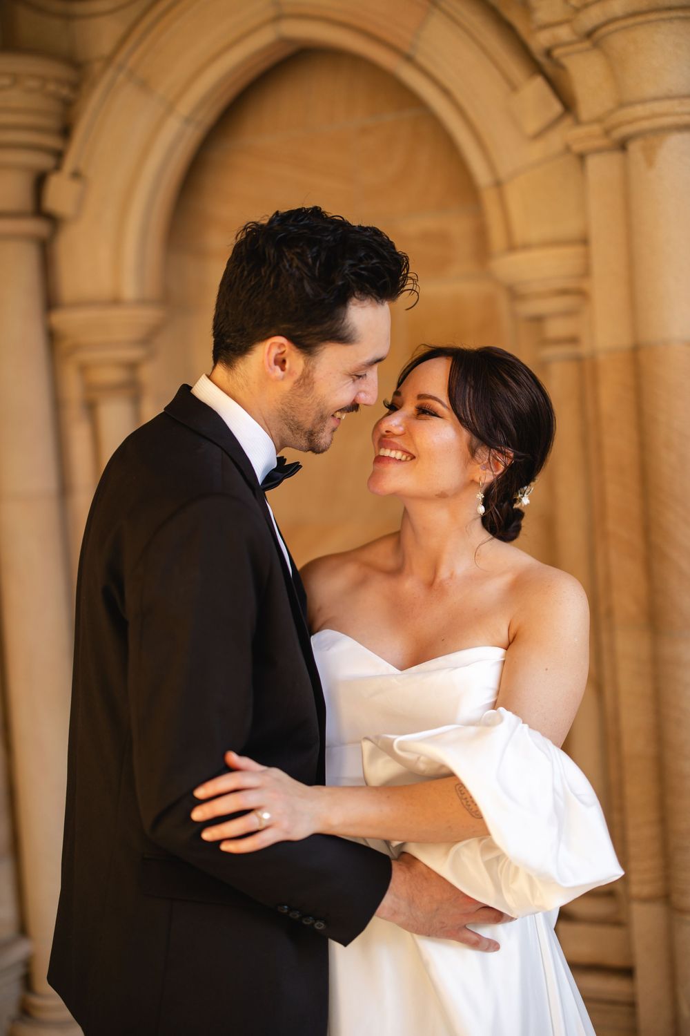 Bride and groom share a joyful embrace beside ornate stone arches in a warm golden light setting.