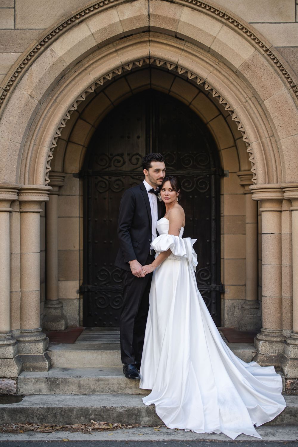 Bride in a flowing white gown and groom in black tuxedo pose beneath a grand Gothic church archway.