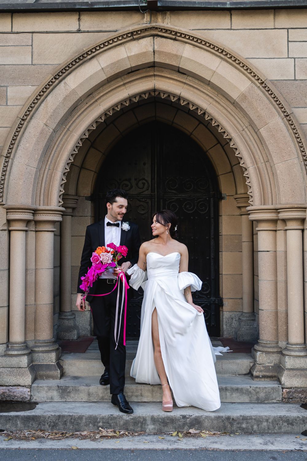 Bride and groom posing at ornate stone church arch doorway, bride in white strapless gown holding vibrant pink bouquet.