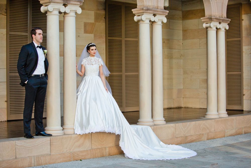 Elegant bride in cathedral-length gown and veil standing beside groom among classical sandstone columns.