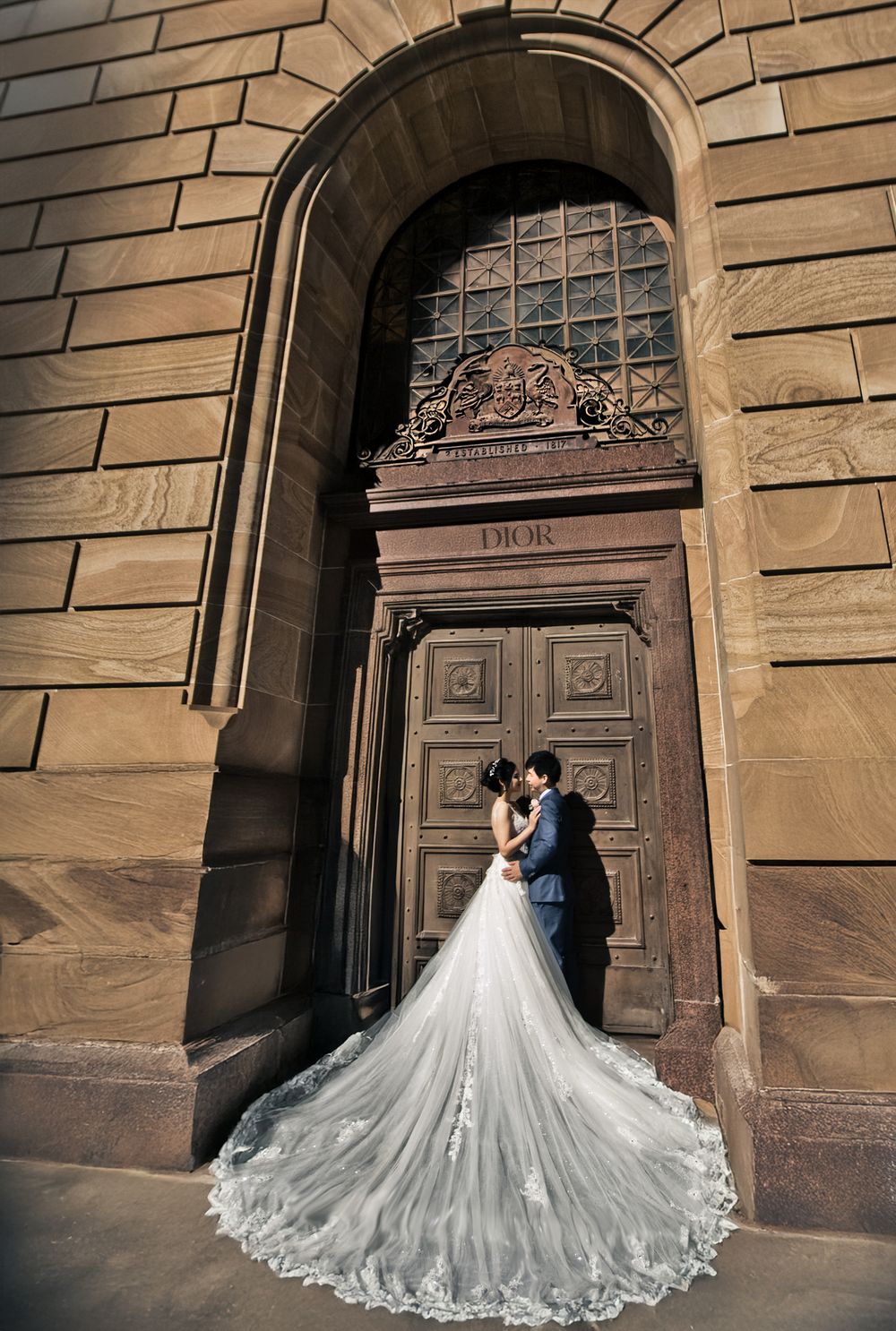 Wedding couple embracing before a grand Dior sandstone archway with sweeping lace cathedral train.
