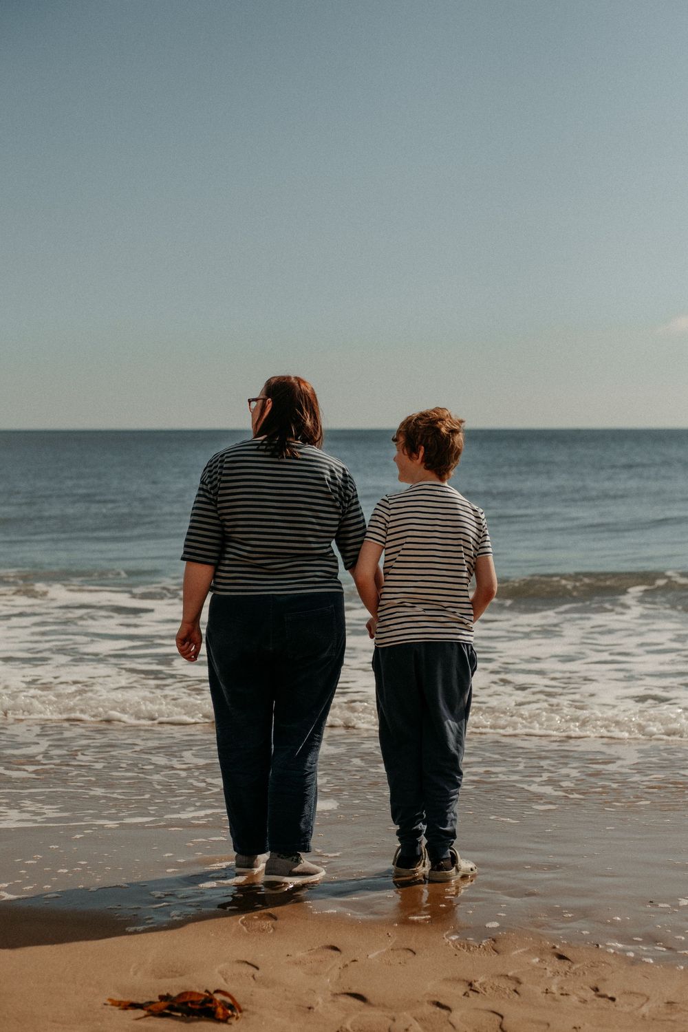 Mother and autistic son standing on the beach looking out to the horizon - their backs facing the viewer. They are wearing matching striped