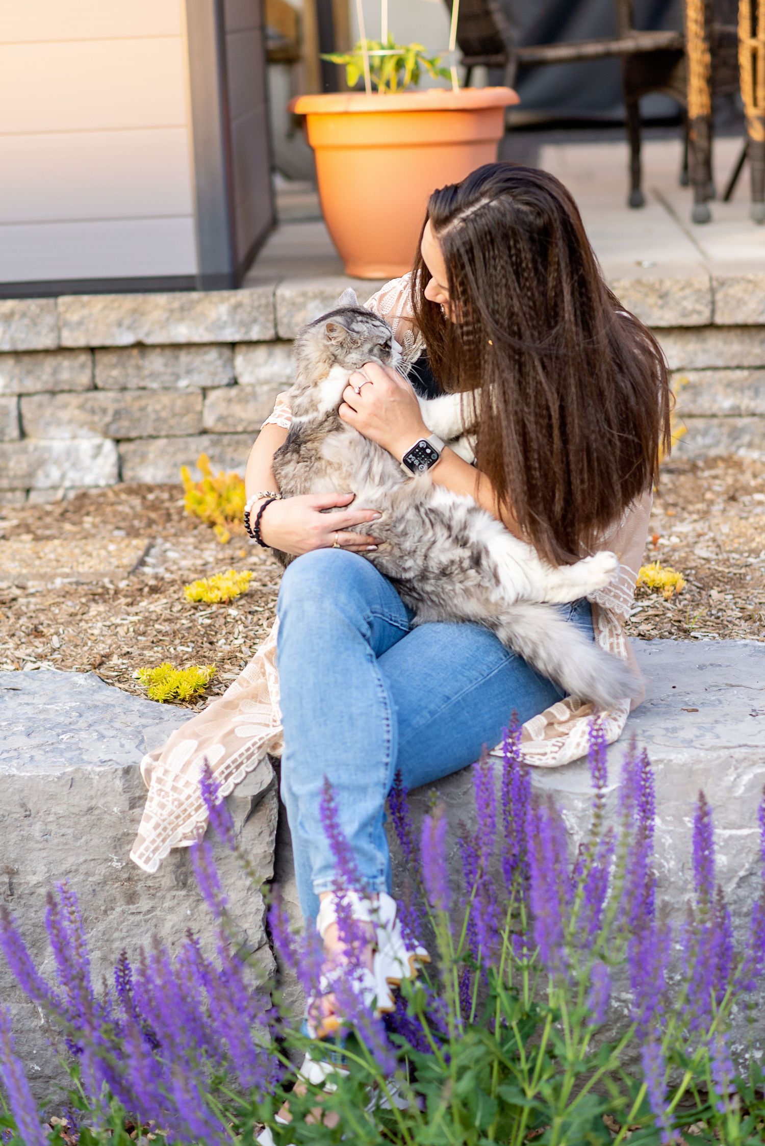 A gray cat cuddles affectionately with someone sitting near purple flowers in a garden setting.