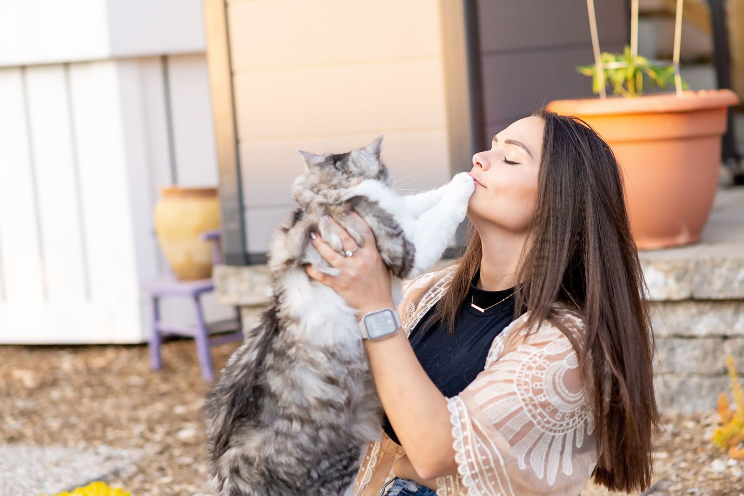 A woman with long dark hair plays with a fluffy grey cat outdoors in a collage of four similar photos.