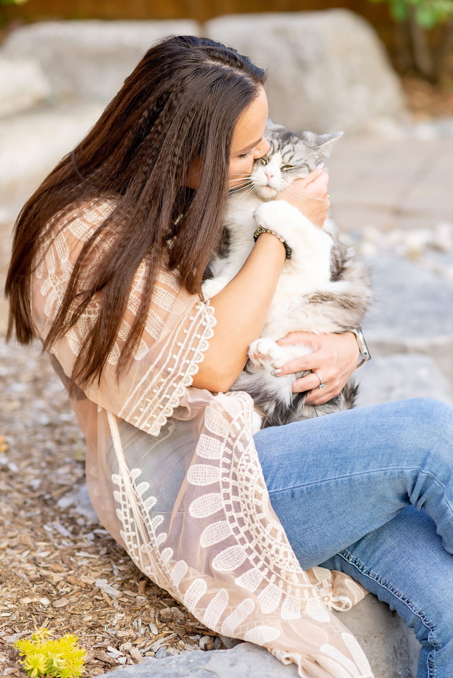 A tabby cat snuggles close to someone wearing a lace top and jeans in an outdoor setting.