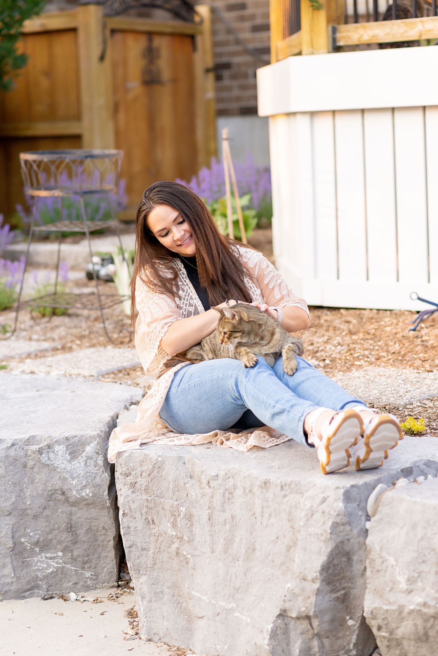 Someone relaxes on stone steps with a cat in a garden setting with a white fence and brick wall.