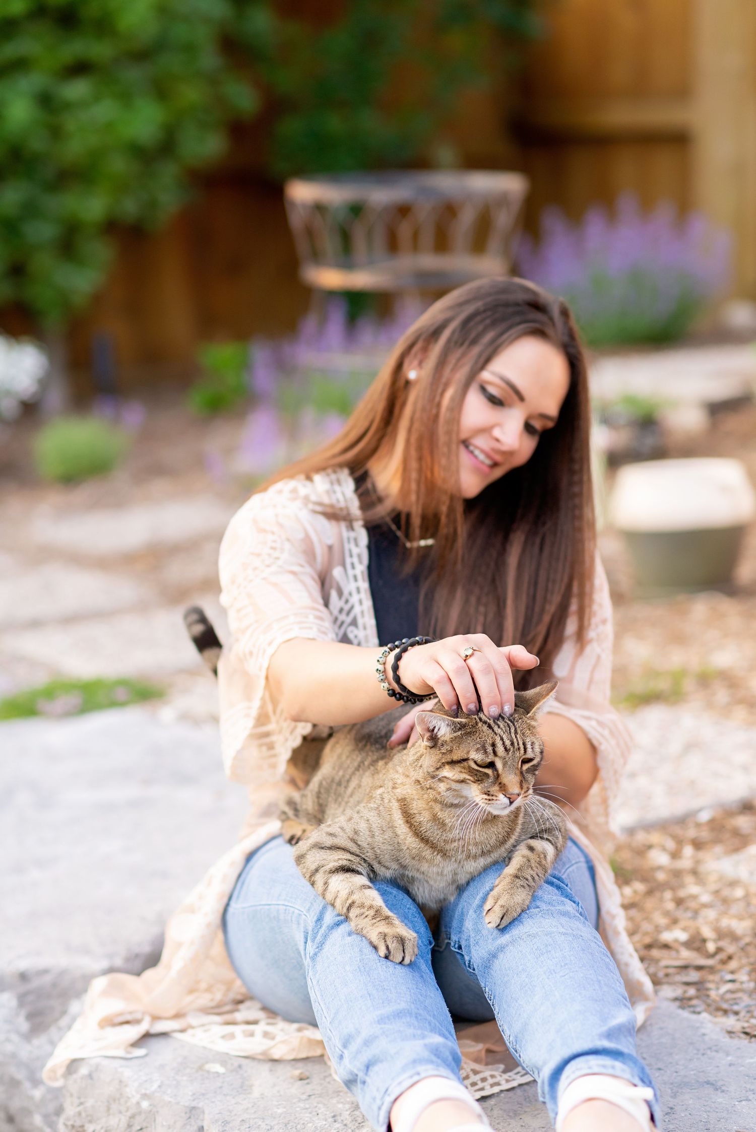 Person in light sweater and jeans lying on ground playing with striped tabby cat outdoors.