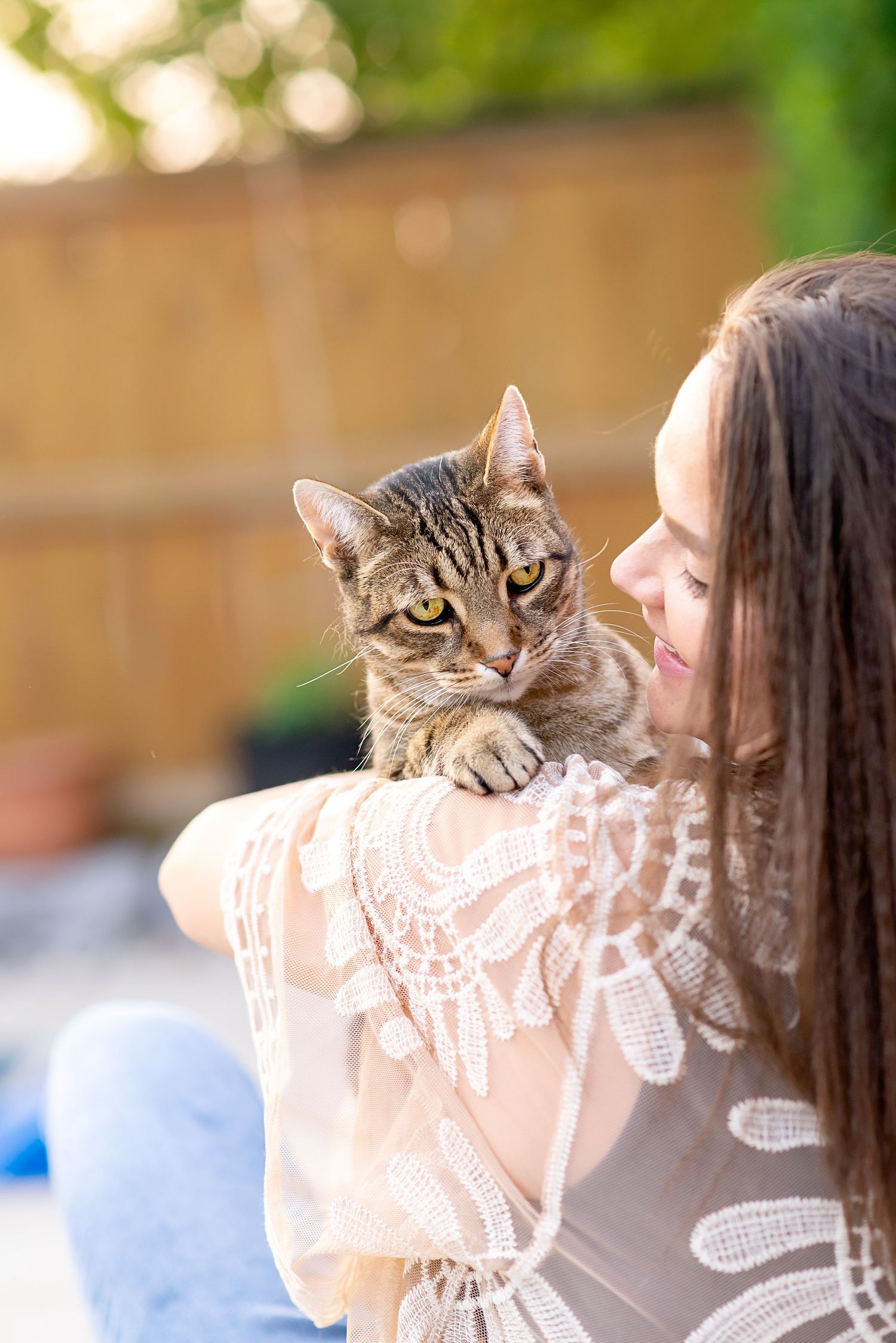 A curious brown tabby cat with bright eyes peeks over a shoulder while being held outdoors.