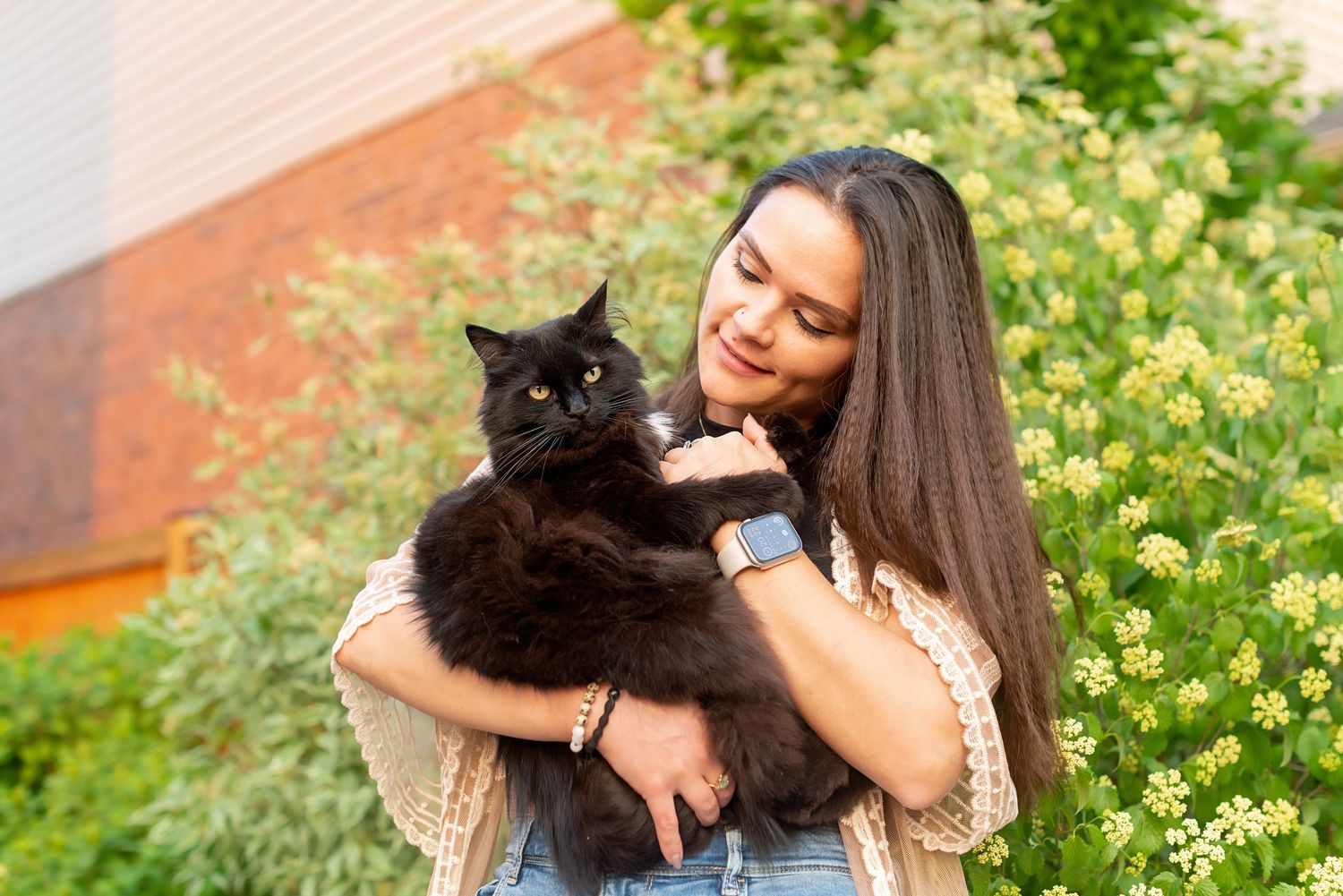 A black cat is held lovingly outdoors against a backdrop of yellow wildflowers and a brick wall.
