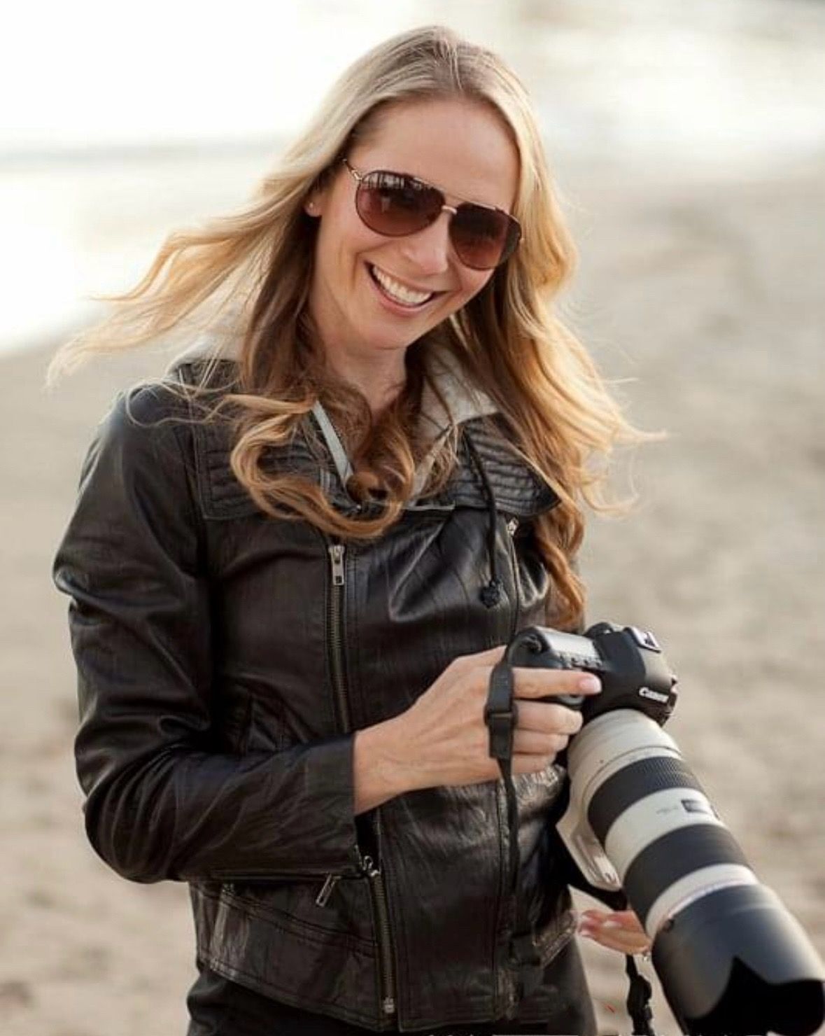 Professional photographer in leather jacket smiles while holding camera with telephoto lens outdoors.