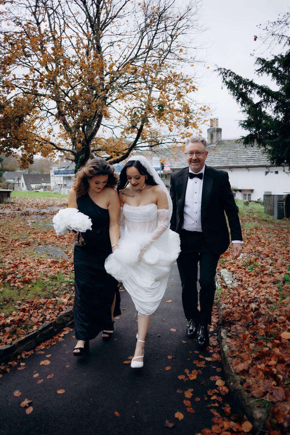 Wedding party walks playfully through autumn leaves on a path with the bride's white dress flowing.