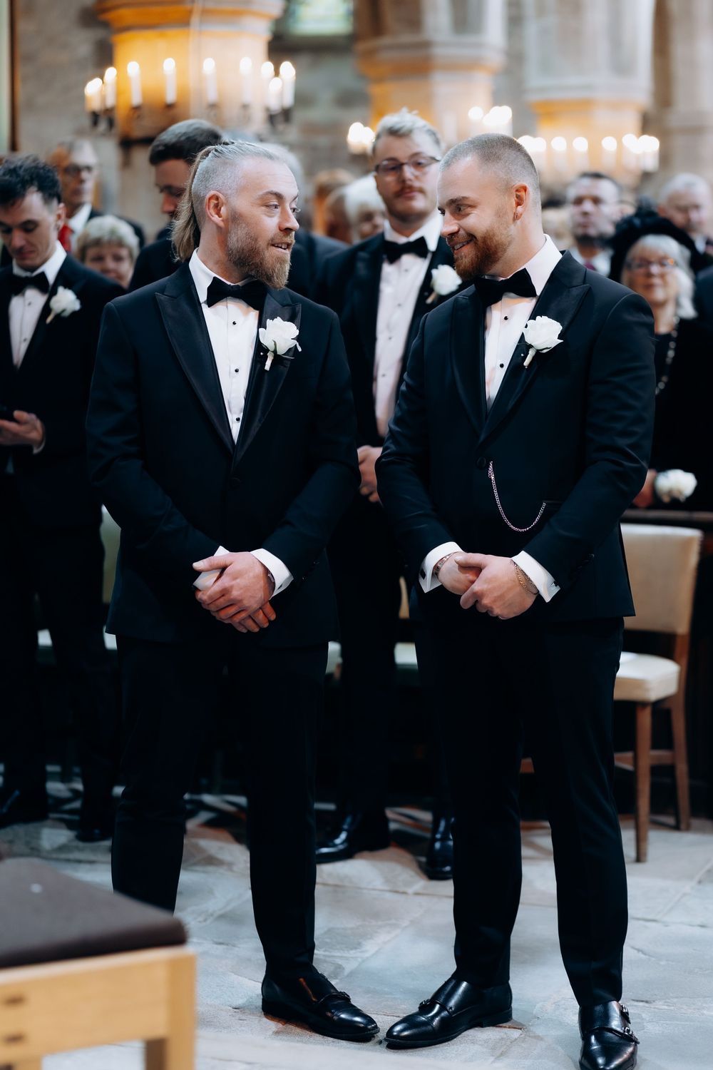 Groomsmen in black tuxedos share a lighthearted moment during a formal wedding ceremony.