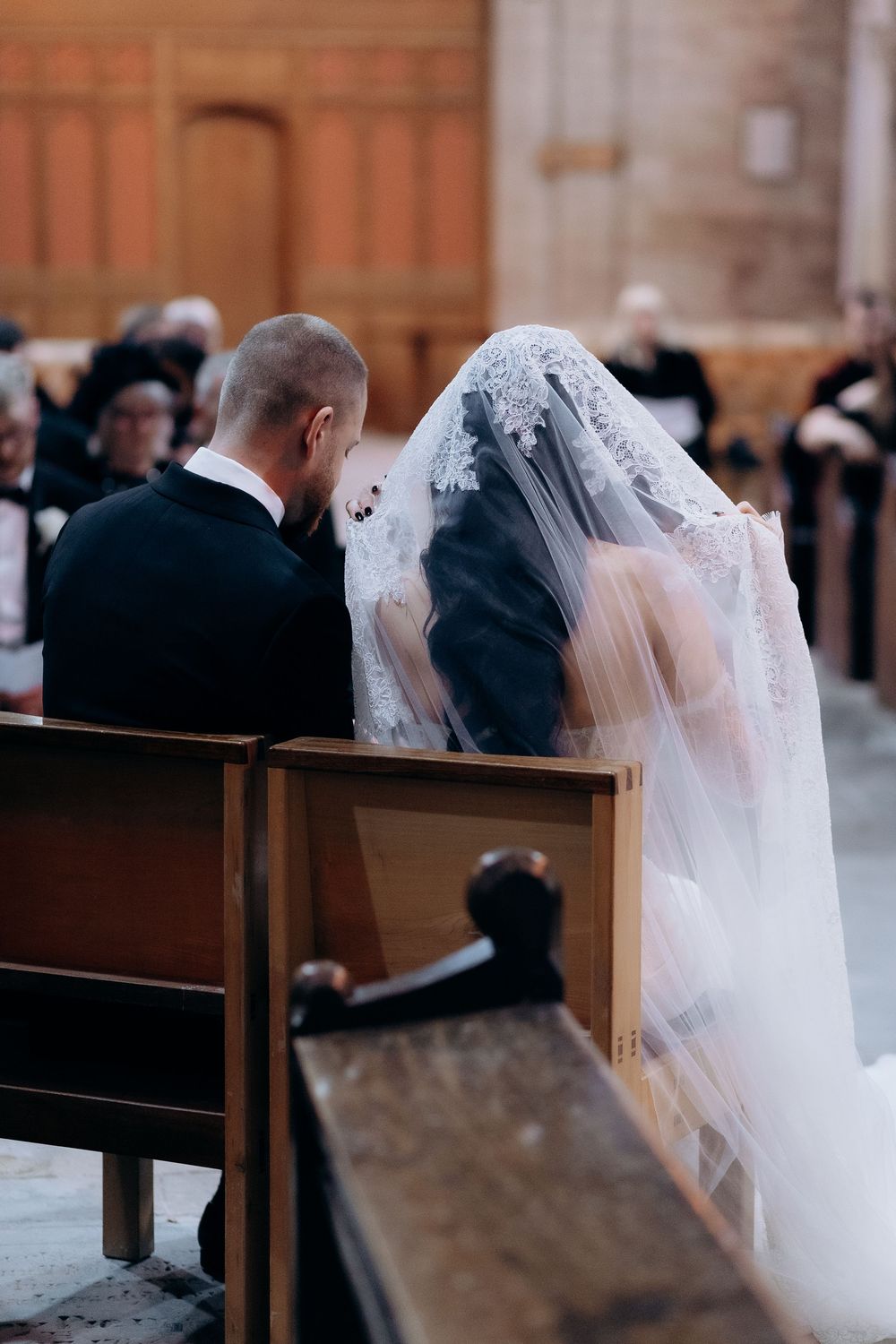 Side view of wedding ceremony attendees seated in wooden church pews with flowing white veil visible.