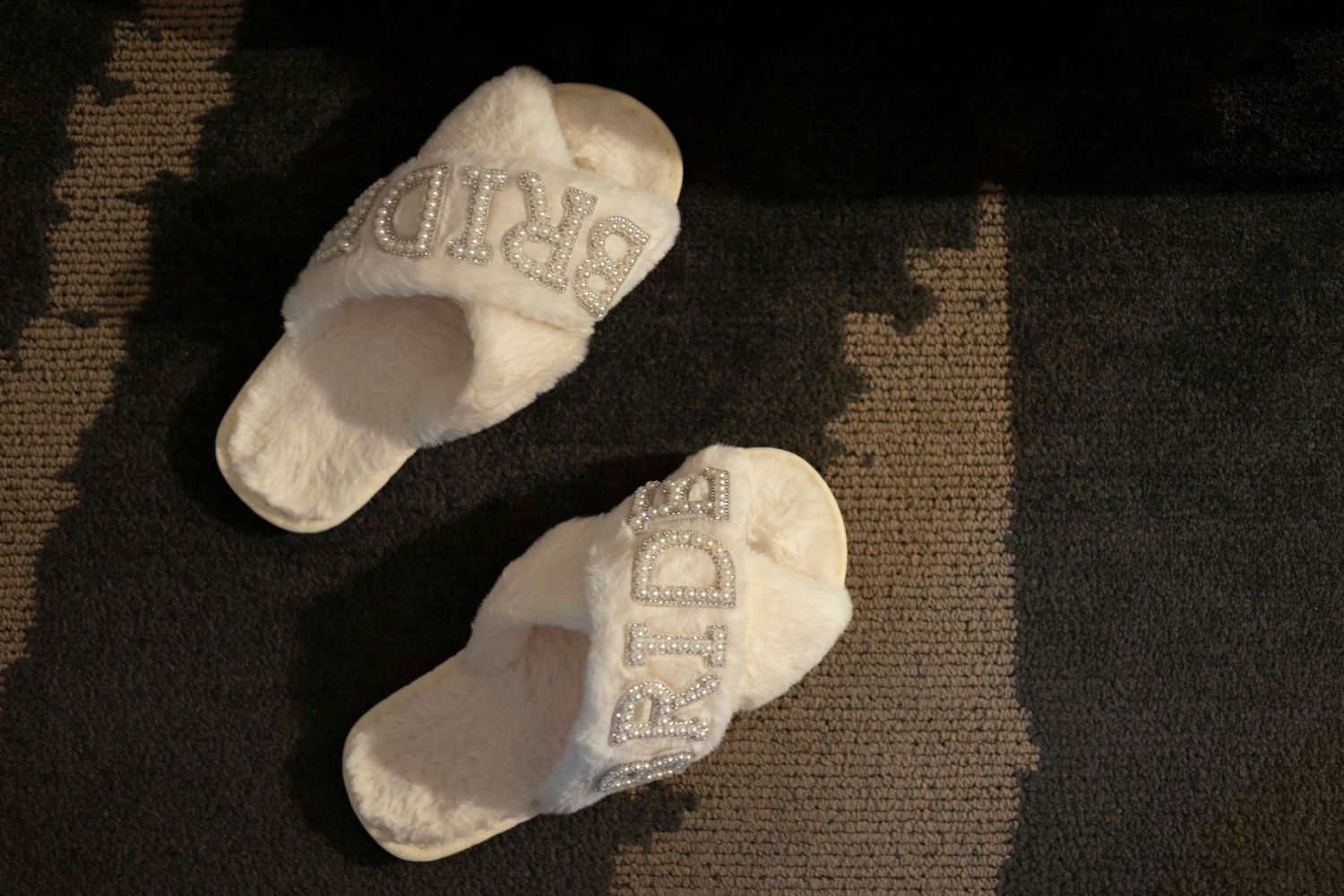 White plush slippers with embroidered text cast shadows on a dark carpet in natural lighting.
